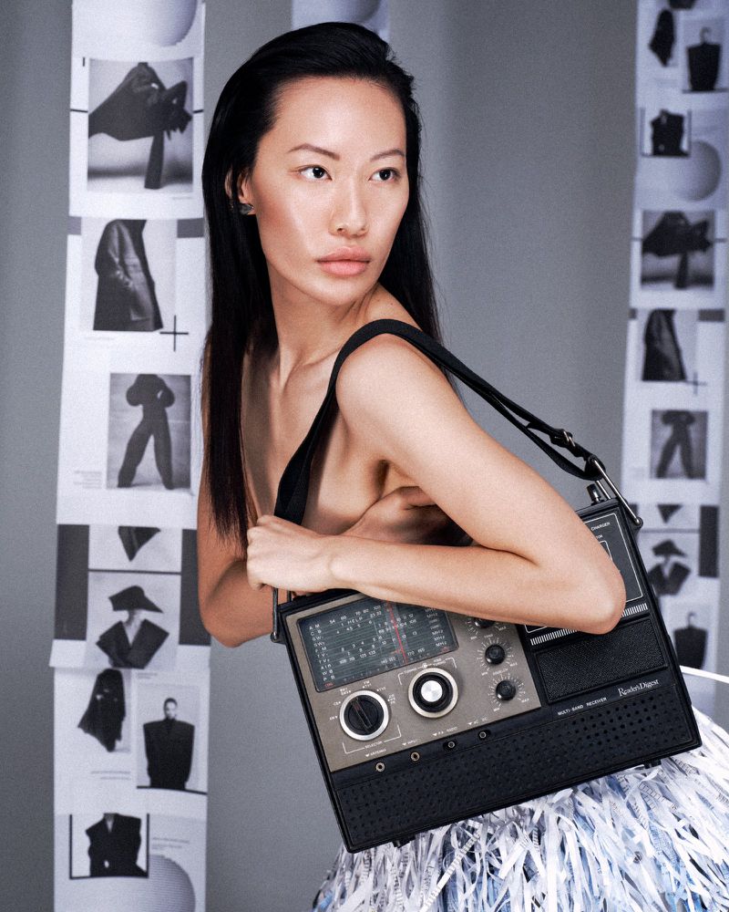 Asian woman in a fashion portrait holding a vintage radio-style handbag, posing against a backdrop of black and white fashion photos.