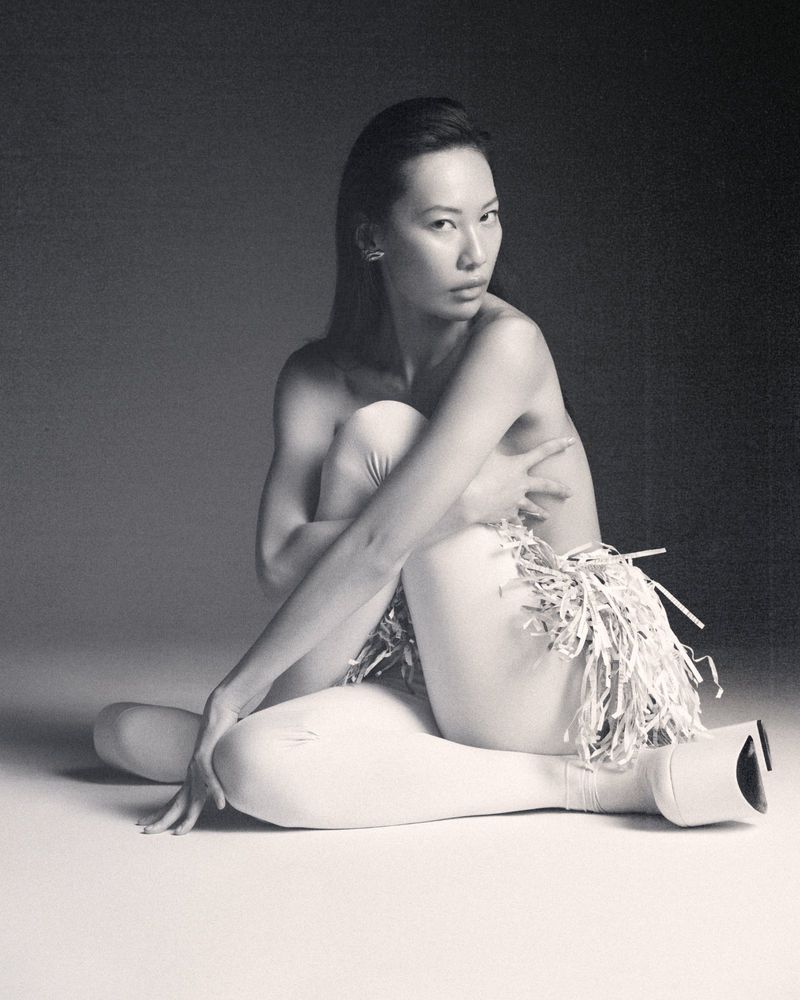Asian woman poses nude in black and white with shredded paper around her waist and high heel boots, in a photo studio setting.