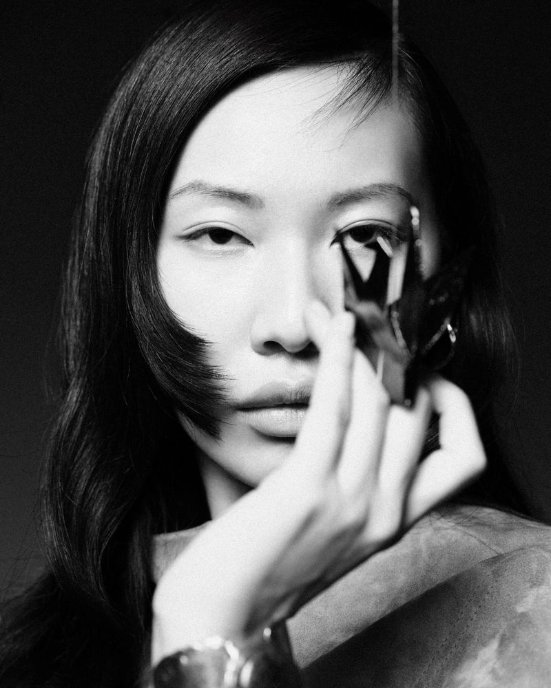 Close-up monochrome portrait of a model holding a dark origami object near her face against a plain dark background.
