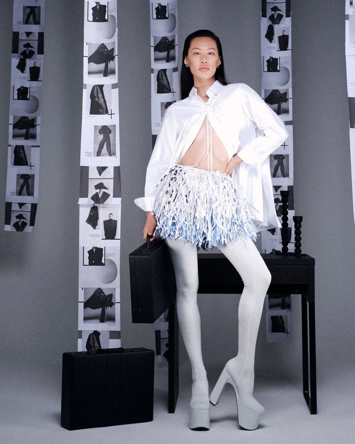 Model Hong Lin posing in an open white shirt, fringe miniskirt, and white platform boots beside black briefcases in a studio setting.