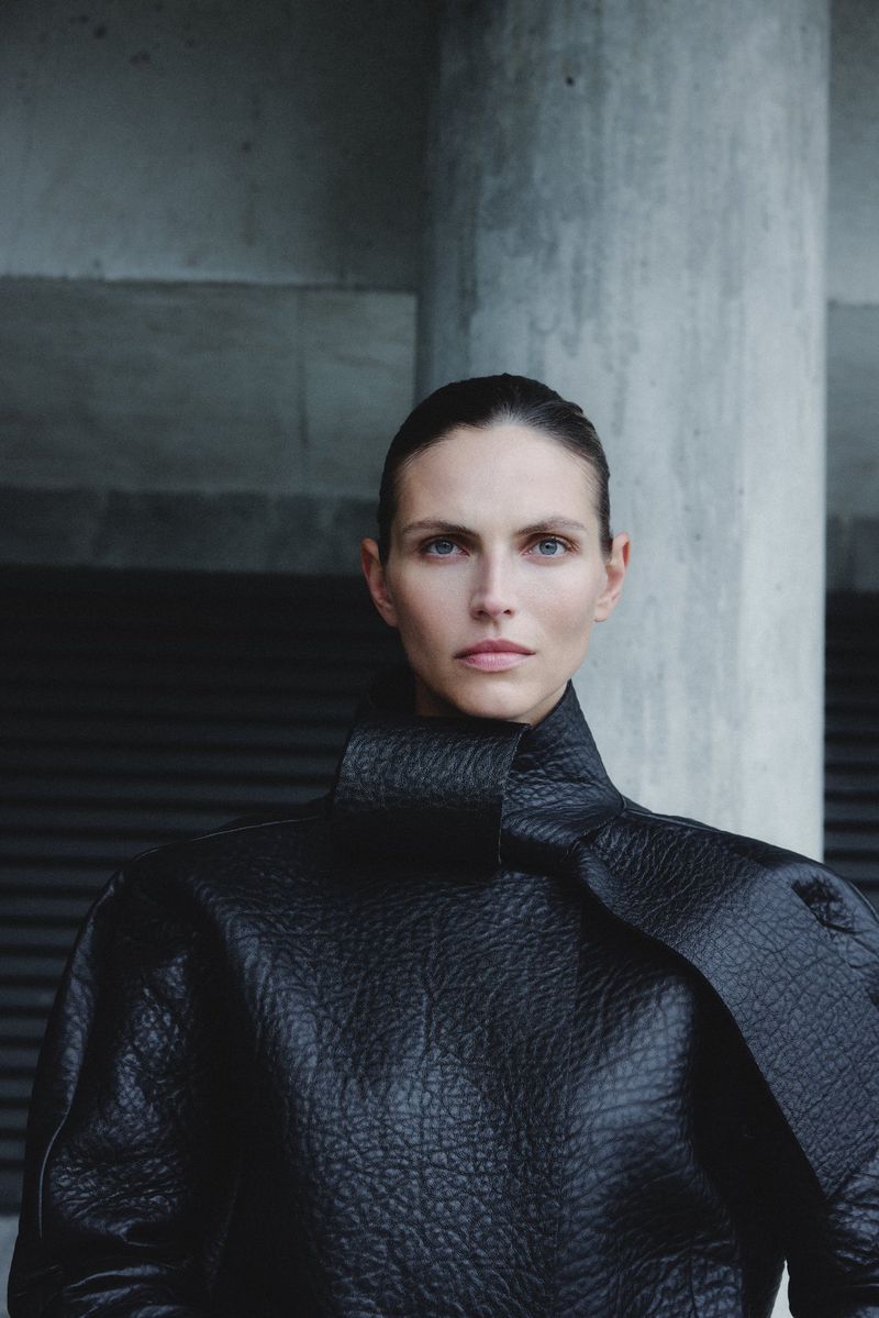 A woman in a black textured coat with a high collar poses against a grey concrete wall. Fashion photography with a neutral expression.
