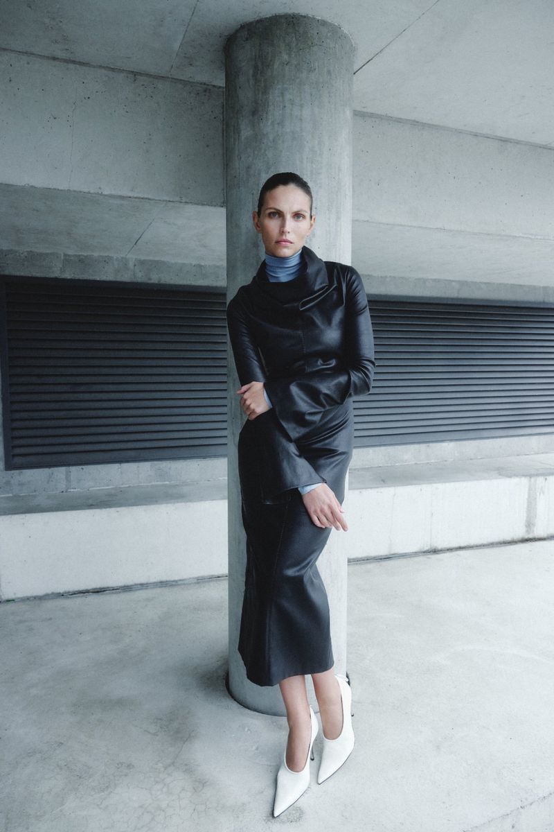 A woman in a black leather dress and white heels poses against a concrete column, her arms crossed, in an urban setting.