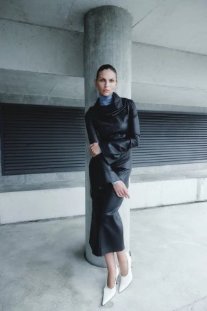 A woman in a black leather dress and white heels poses against a concrete column, her arms crossed, in an urban setting.
