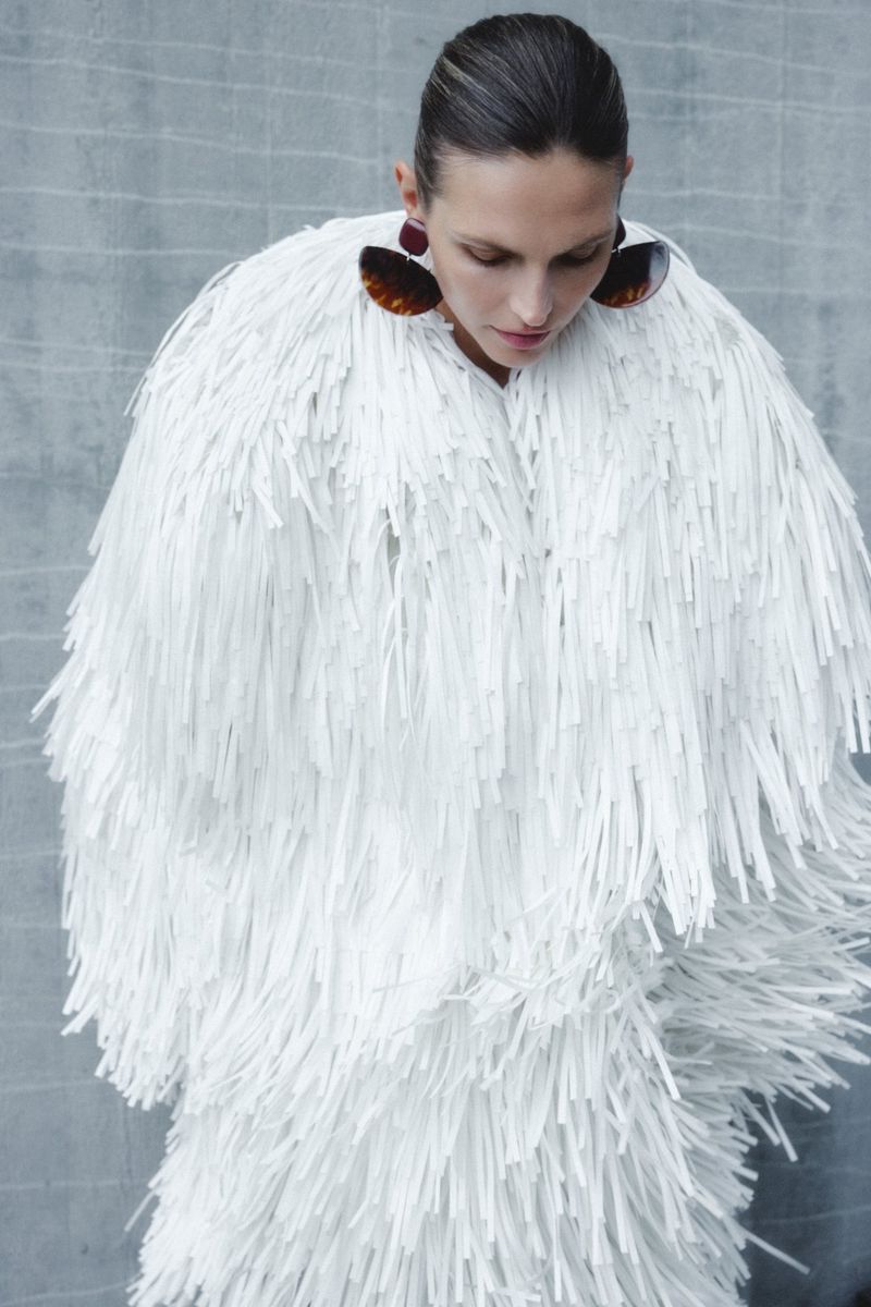 Woman in a white fringed outfit and earrings against a concrete-like background, fashion, style, clothing, and portrait photography.