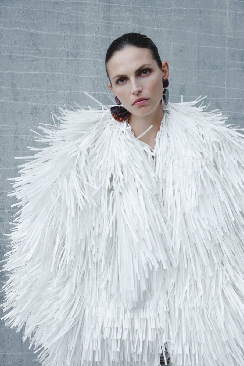 Woman in white fringe jacket poses against gray wall, wearing statement earrings for fashion or beauty editorial shot.