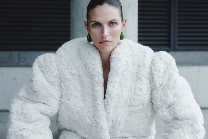 A stylish adult woman wearing a white fur coat and green earrings, posing in an urban setting with a neutral facial expression.
