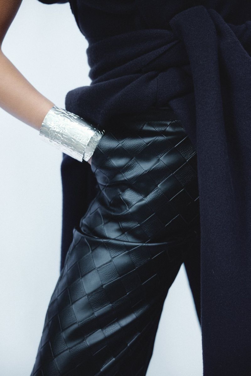 A model is pictured wearing a silver bracelet, black top, and patterned black pants against a plain white backdrop in a studio shot.