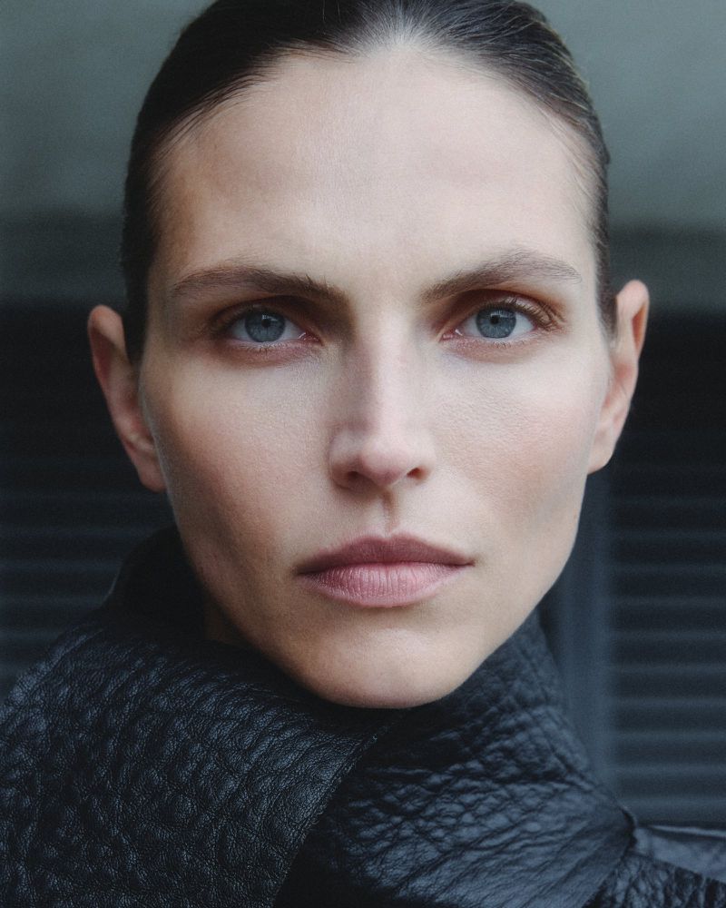 Close-up portrait of a woman with light skin and blue eyes wearing a black jacket against a dark background, captured in natural lighting.