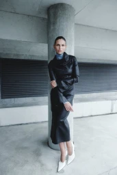 Model Karlina Caune leans against a concrete pillar wearing a black leather ensemble and white pointed-toe heels.