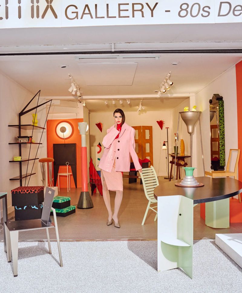 Woman in pink suit in a modern art gallery with geometric furniture, bright lighting, and Memphis design elements, showcasing 80s decor.