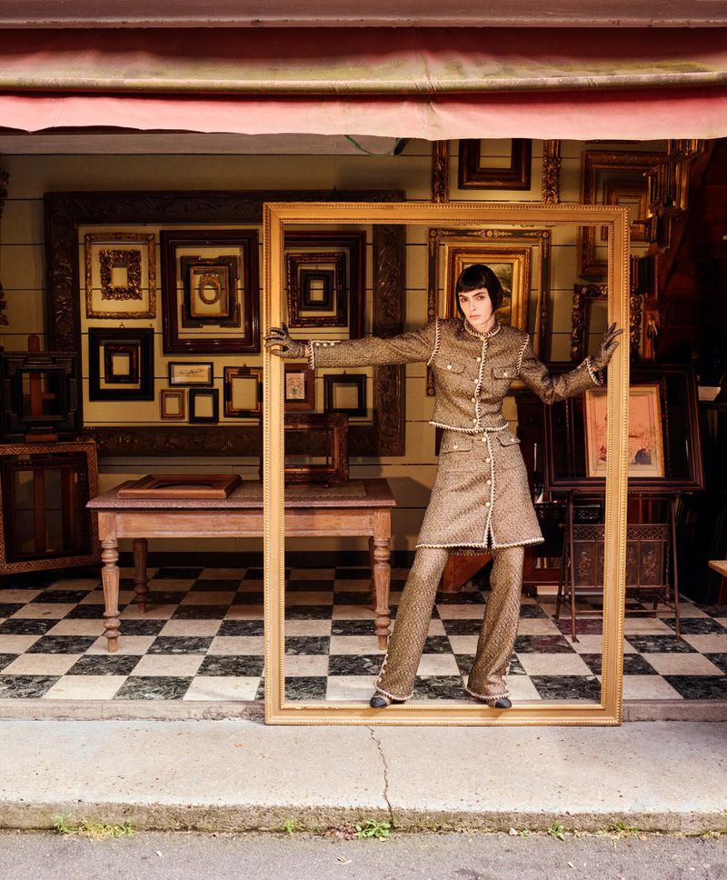 Fashion model posing in tweed pantsuit, framed by a large gold picture frame outside an art gallery with checkered tile floor.