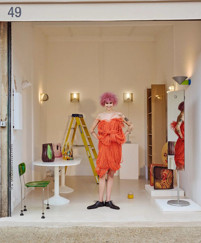 Woman with pink hair in a textured orange dress and black shoes stands in a white interior space with a table and ladder.