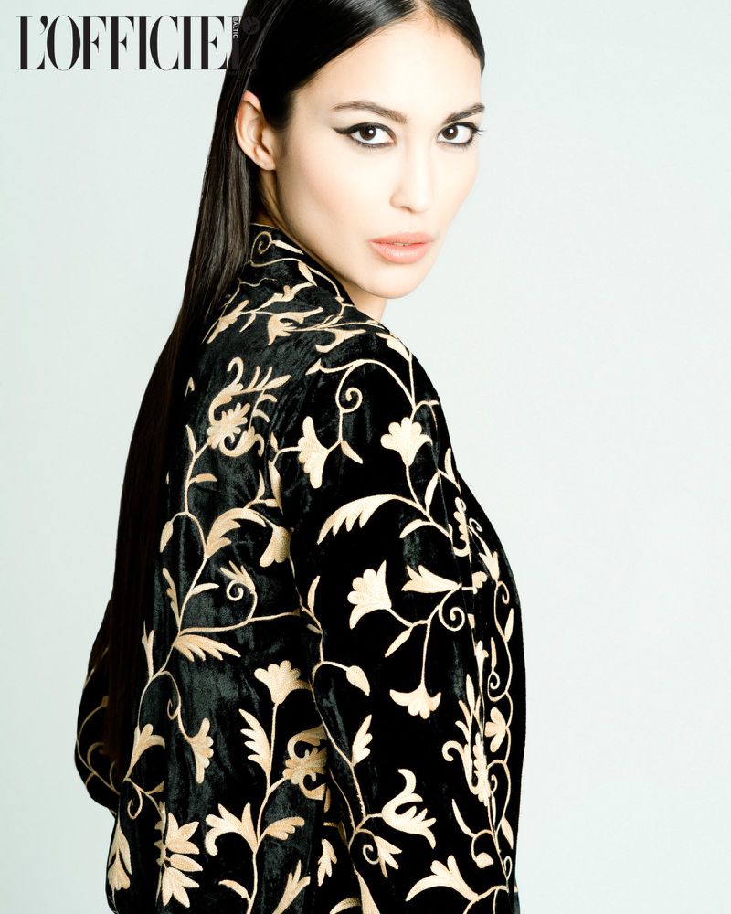 Portrait of a woman with dark hair wearing a black velvet jacket with gold floral embroidery against a light grey background.