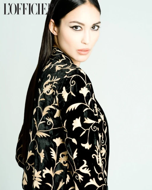 Portrait of a woman with dark hair wearing a black velvet jacket with gold floral embroidery against a light grey background.