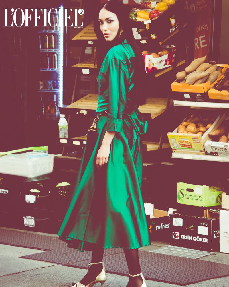 Fashion editorial shot of a woman in a green satin dress posing in front of a grocery store display, with the magazine logo visible.