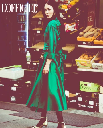 Fashion editorial shot of a woman in a green satin dress posing in front of a grocery store display, with the magazine logo visible.