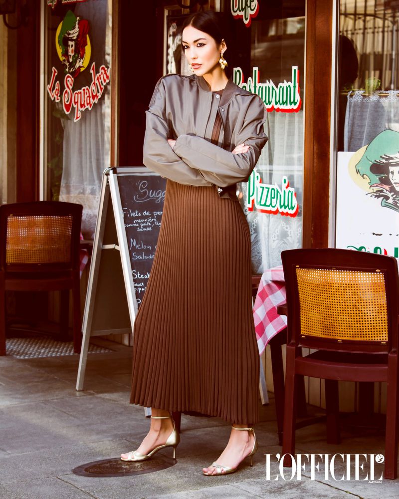 A fashionable woman poses in front of a pizzeria wearing a grey jacket, pleated brown skirt, and gold heels in an urban setting.