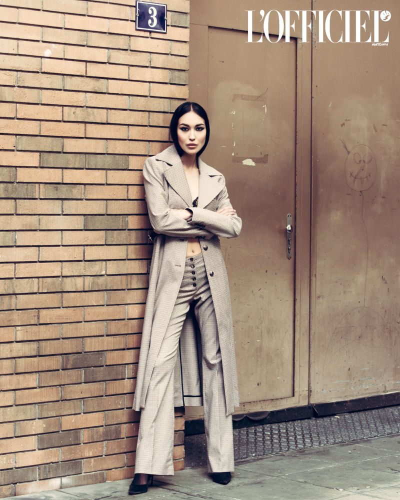 Fashion editorial shot of a slender woman in a trench coat and pantsuit posing against a brick wall and metal door in an urban setting.