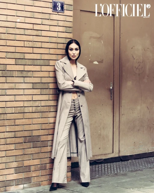 Fashion editorial shot of a slender woman in a trench coat and pantsuit posing against a brick wall and metal door in an urban setting.