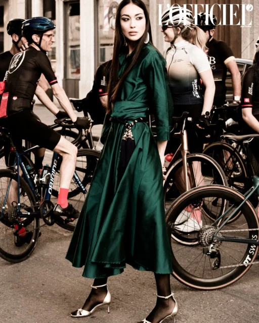 Woman in green dress poses in front of group of cyclists on street with bicycles and European architecture, editorial fashion shot.