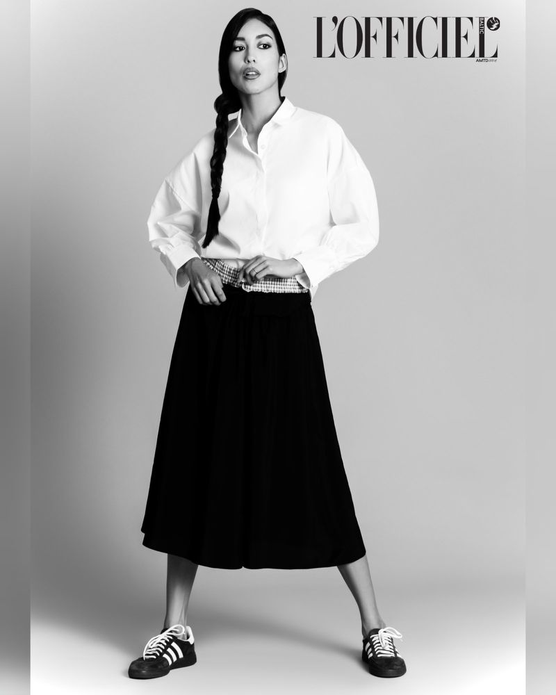 Black-and-white full shot of a woman in a white button-down shirt and black skirt, posing in sneakers with the magazine title L'Officiel.