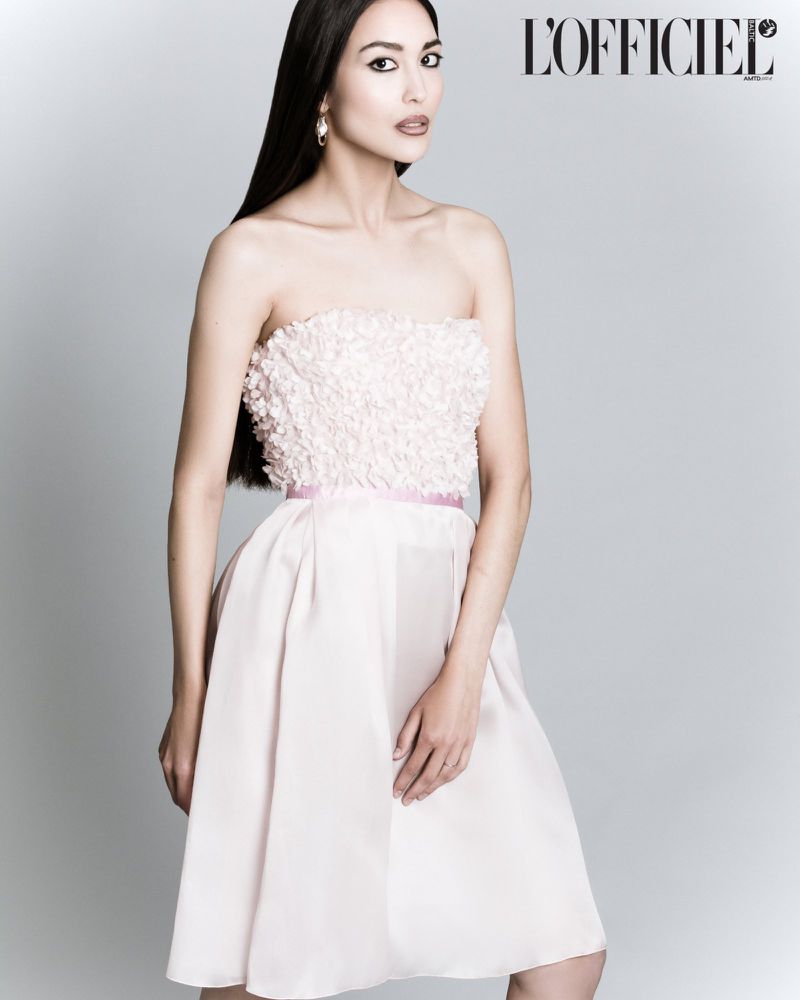 Elegant woman in a light pink strapless dress posing in a studio, long dark hair, minimalist earrings, light grey background.