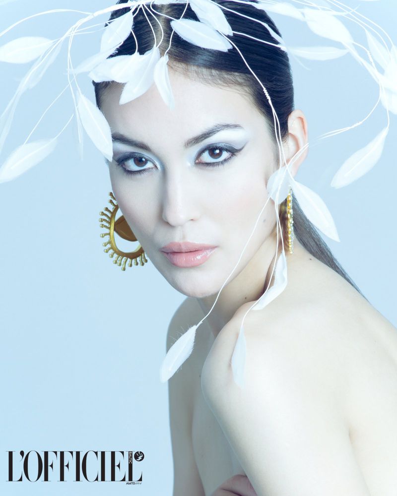 Close-up studio shot of a beautiful adult woman with dramatic makeup, statement earrings, and a feather headpiece on a pale-blue background.