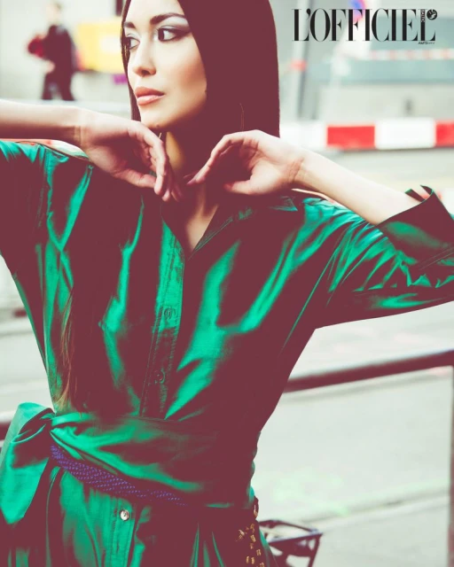 Woman in green dress with purple belt gazes to the side, hands near her face in an outdoor fashion editorial for L'Officiel magazine.