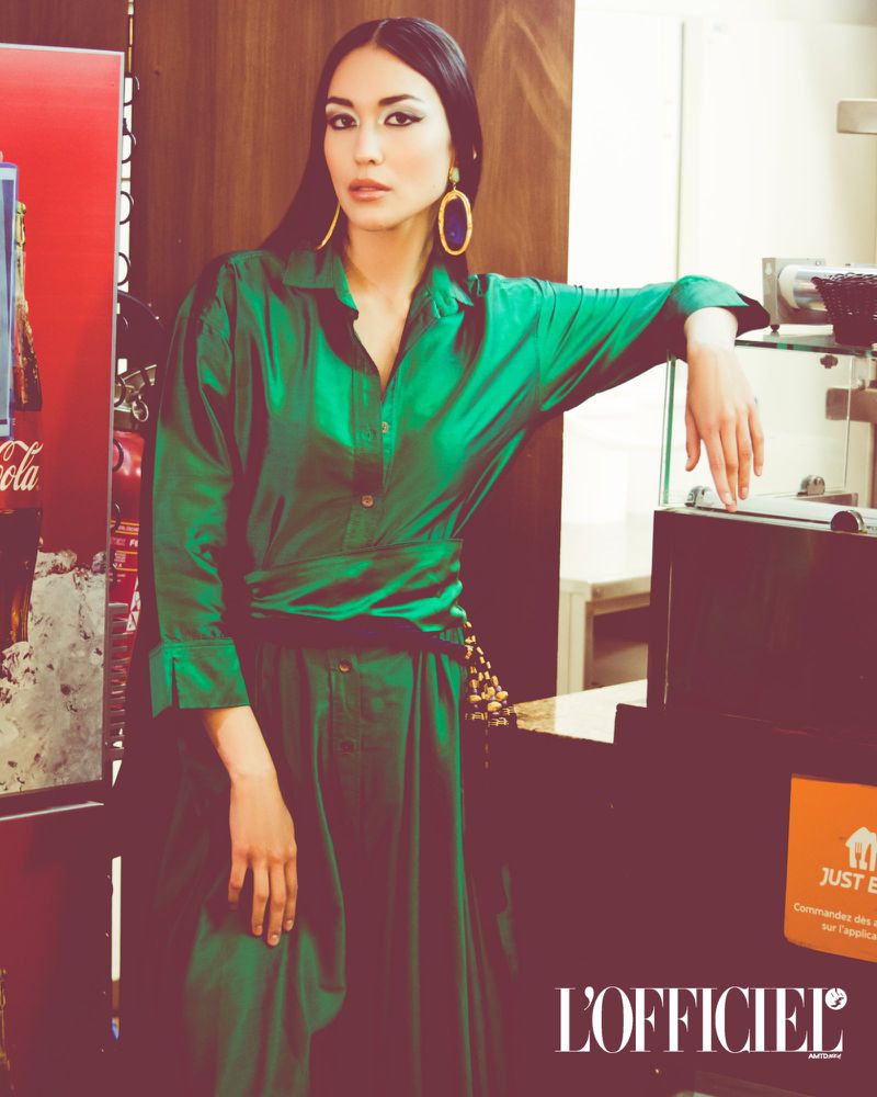Brunette woman in a shiny green dress and large hoop earrings leans against a vending machine in an indoor environment.
