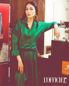 Woman wearing a bright emerald green silk shirt dress and large drop earrings leaning against a counter.