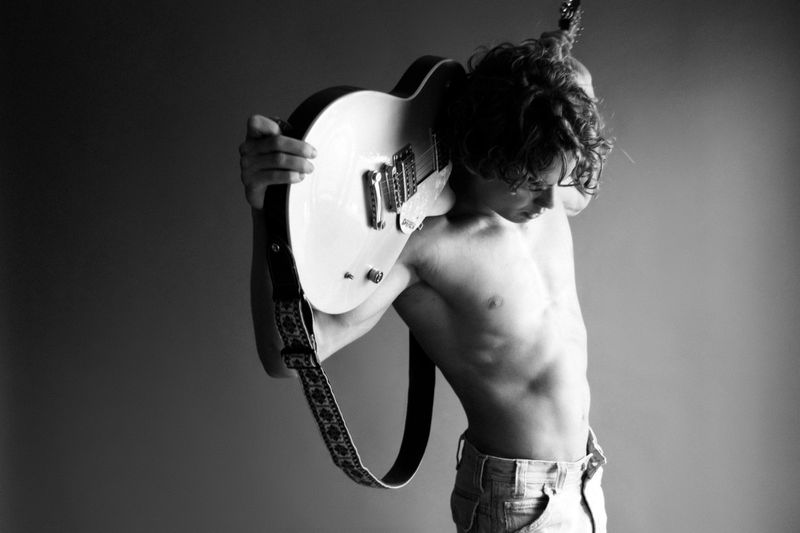 Black-and-white studio portrait of a shirtless male model holding a guitar over his shoulder, looking down against a grey background.