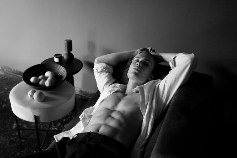 Black and white portrait of a fair-skinned, muscular adult male reclining shirtless on a couch in a low-lit indoor environment.