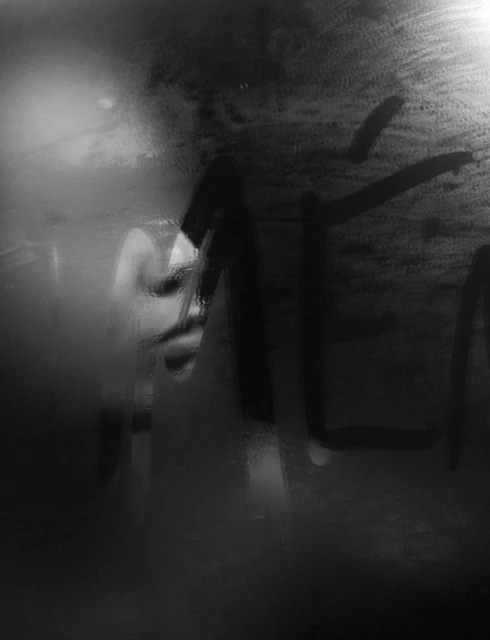 Monochrome close-up of a person's face partially visible behind condensation and abstract writing on glass, creating a hazy effect.