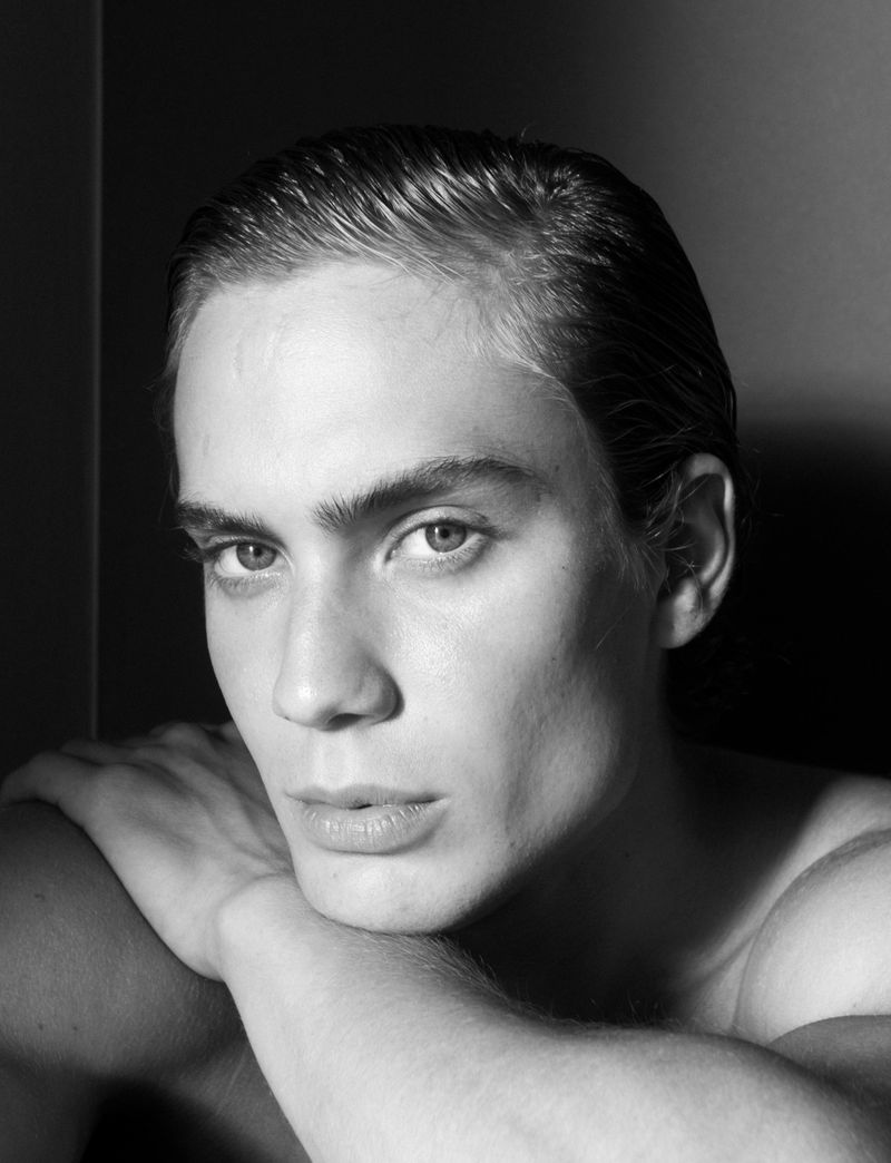 Black and white close-up portrait of a young man with slicked-back hair resting his chin on crossed arms against a dark background.