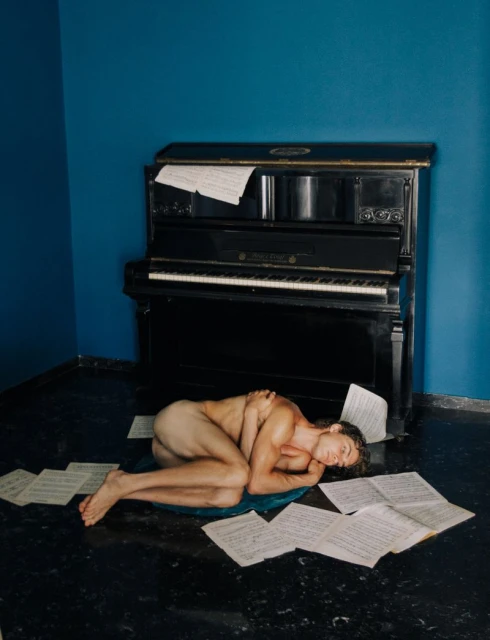 Nude man lies on a cushion near a black piano surrounded by scattered sheet music, set against a dark blue corner wall.