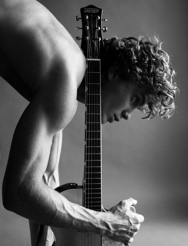 A shirtless, muscular man with curly hair stands indoors holding a Gretsch guitar, captured in a black-and-white studio portrait.