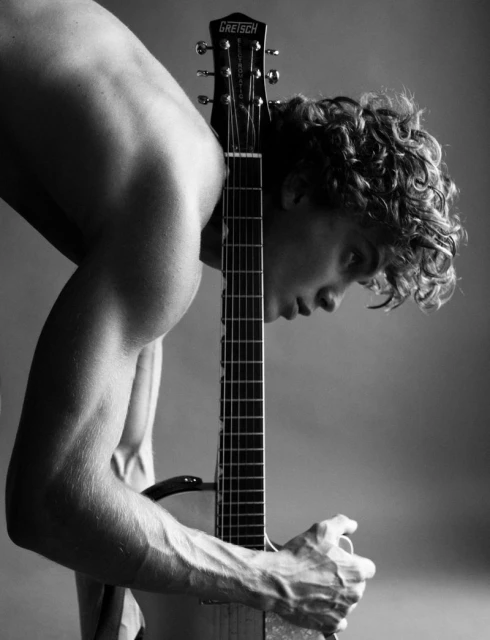A shirtless, muscular man with curly hair stands indoors holding a Gretsch guitar, captured in a black-and-white studio portrait.
