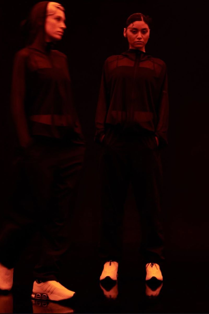 Two models wearing dark hooded jackets and white sneakers posing in a dark studio illuminated by warm red lighting.