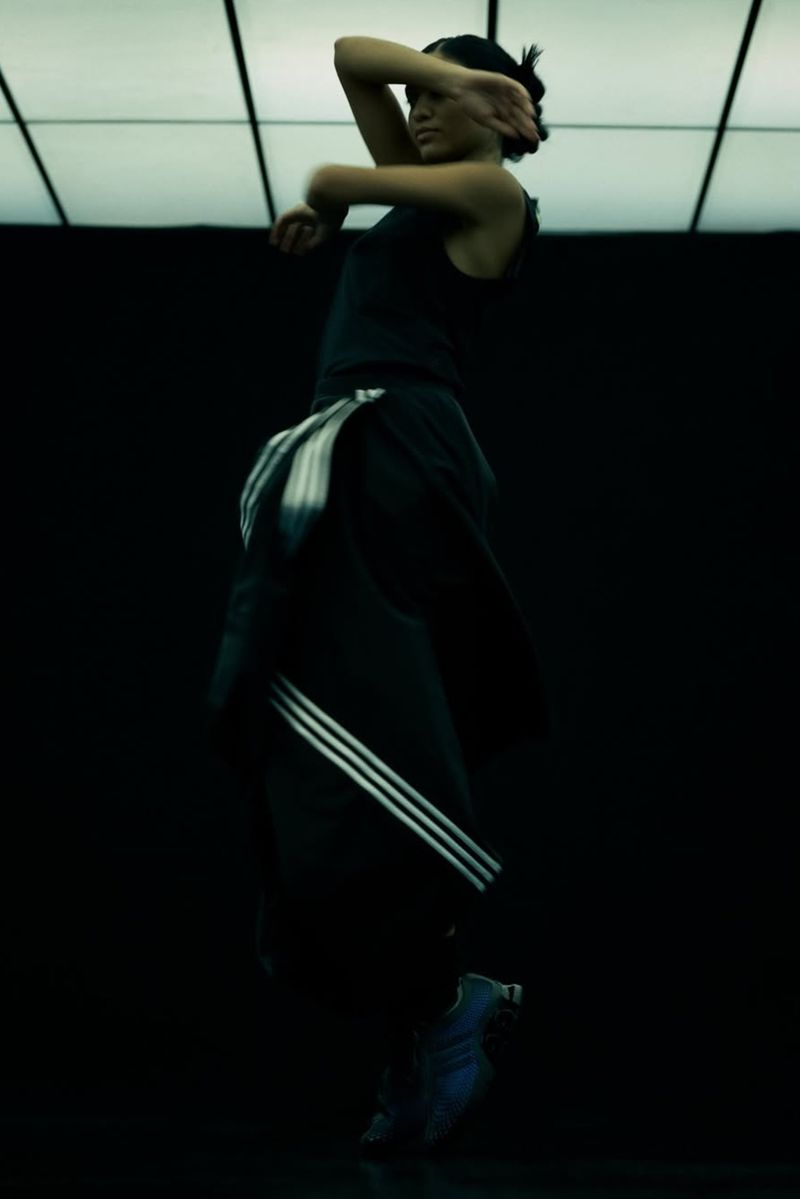 Athletic adult female in sports clothing performs a dance movement in a dimly lit studio space with tiled lighting above.