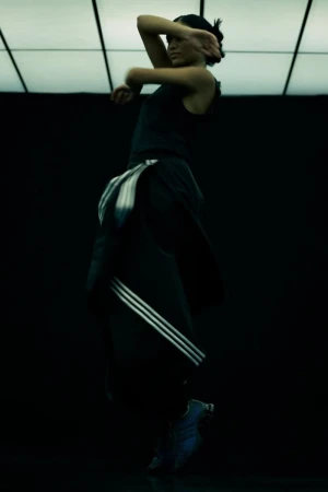 Athletic adult female in sports clothing performs a dance movement in a dimly lit studio space with tiled lighting above.