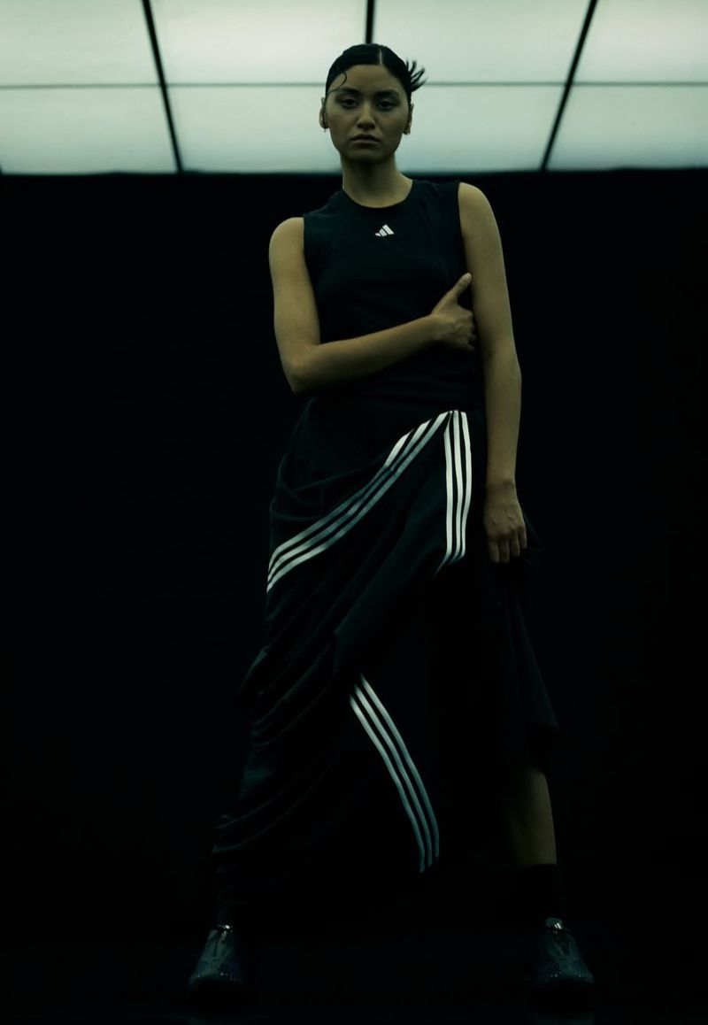 Stylish woman poses in Adidas tank top and skirt with signature 3-stripes, against a dark backdrop with modern, geometric overhead lighting.