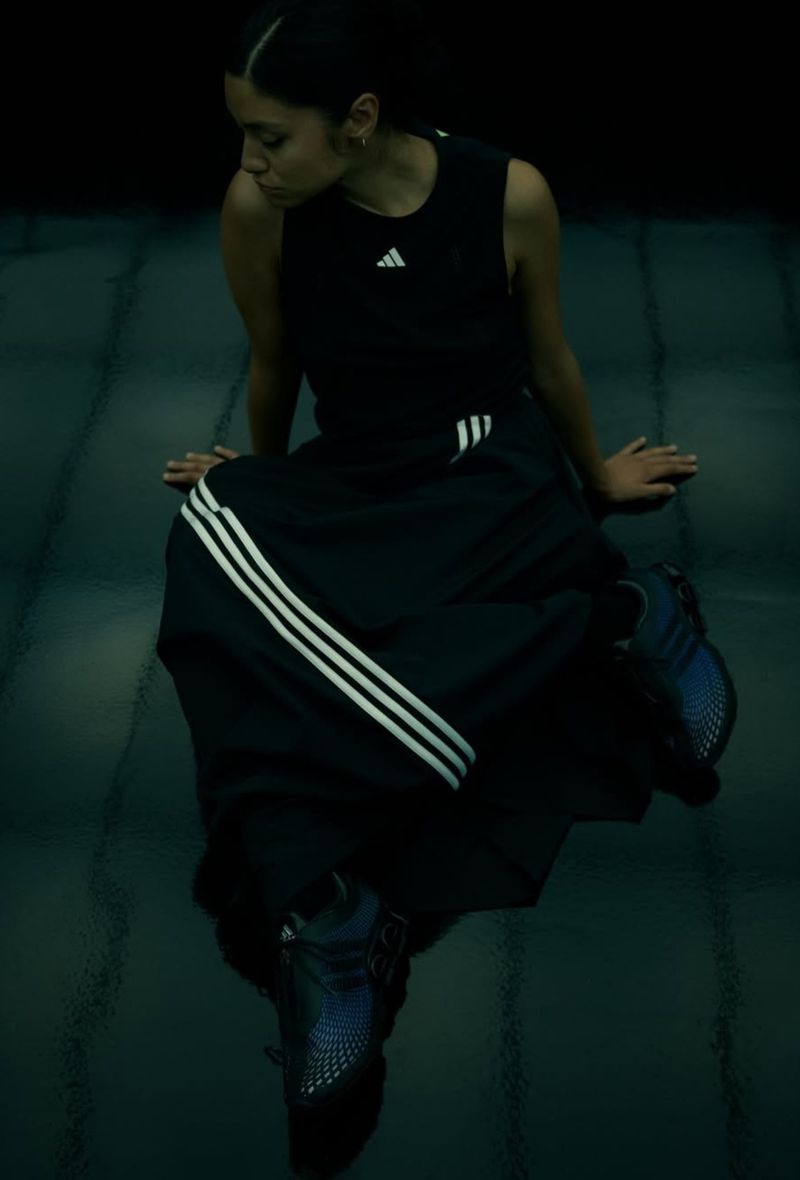 Woman in black athletic wear, including a tank top and loose pants with white stripes, sits indoors with Adidas sneakers and a reflective floor.