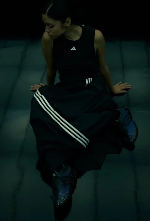 Woman in black athletic wear, including a tank top and loose pants with white stripes, sits indoors with Adidas sneakers and a reflective floor.