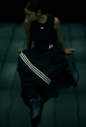 Model seated on a dark reflective floor wearing a black sleeveless top, three-stripe maxi skirt, and sneakers for Adidas x Packer.