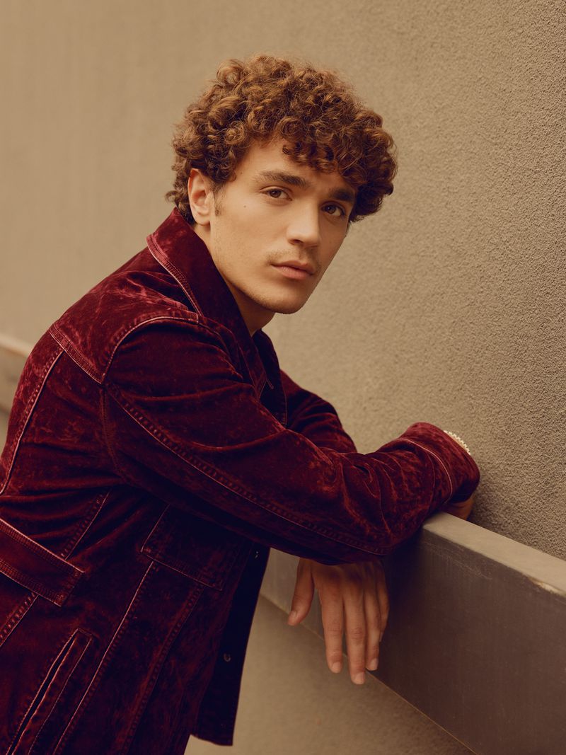 Young man with curly hair in a velvet jacket leans against a ledge indoors. Moody lighting and earth tones evoke timeless style.