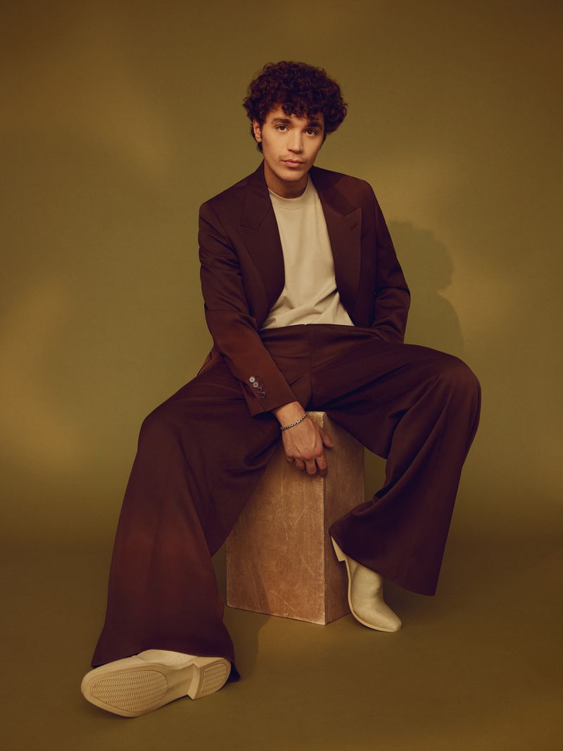 Young adult male in a brown suit sits on a box in a studio. He wears white boots and his curly hair is prominent against the green backdrop.