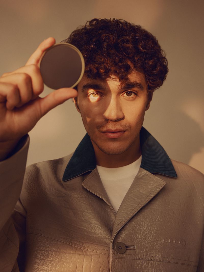 Close-up portrait of a young man with curly hair holding a round compact mirror in front of his eye, wearing a stylish tan jacket.