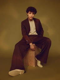 David Iacono seated on a wooden cube wearing an oversized brown suit and cream boots against an olive green backdrop.