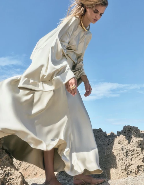 Woman in a flowing beige dress and gold collar poses on rocky ground against a clear blue sky in a fashion editorial photograph.