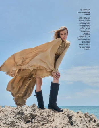 A woman in a flowing gold dress and black boots poses on rocks near the ocean under a clear sky for a fashion editorial.